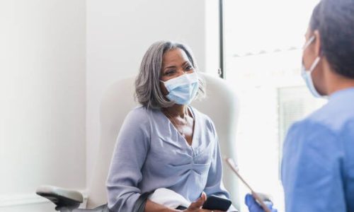 A senior woman holds attentively listens as a female doctor discusses her health. The patient is holding a smartphone. The patient and doctor are wearing protective face masks.